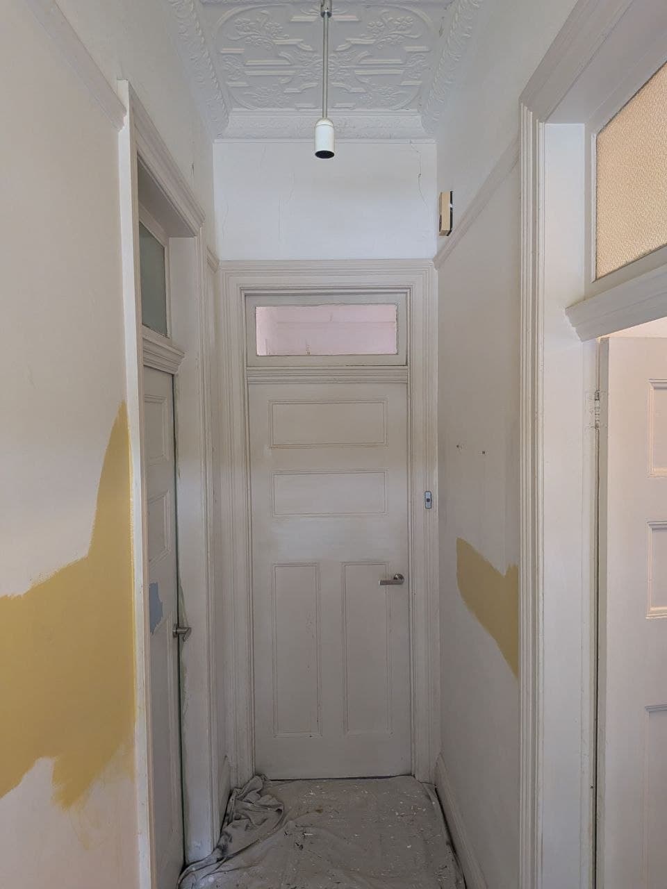 Heritage hallway before repainting — patchy primed walls, bare trim and a drop sheet on the floor