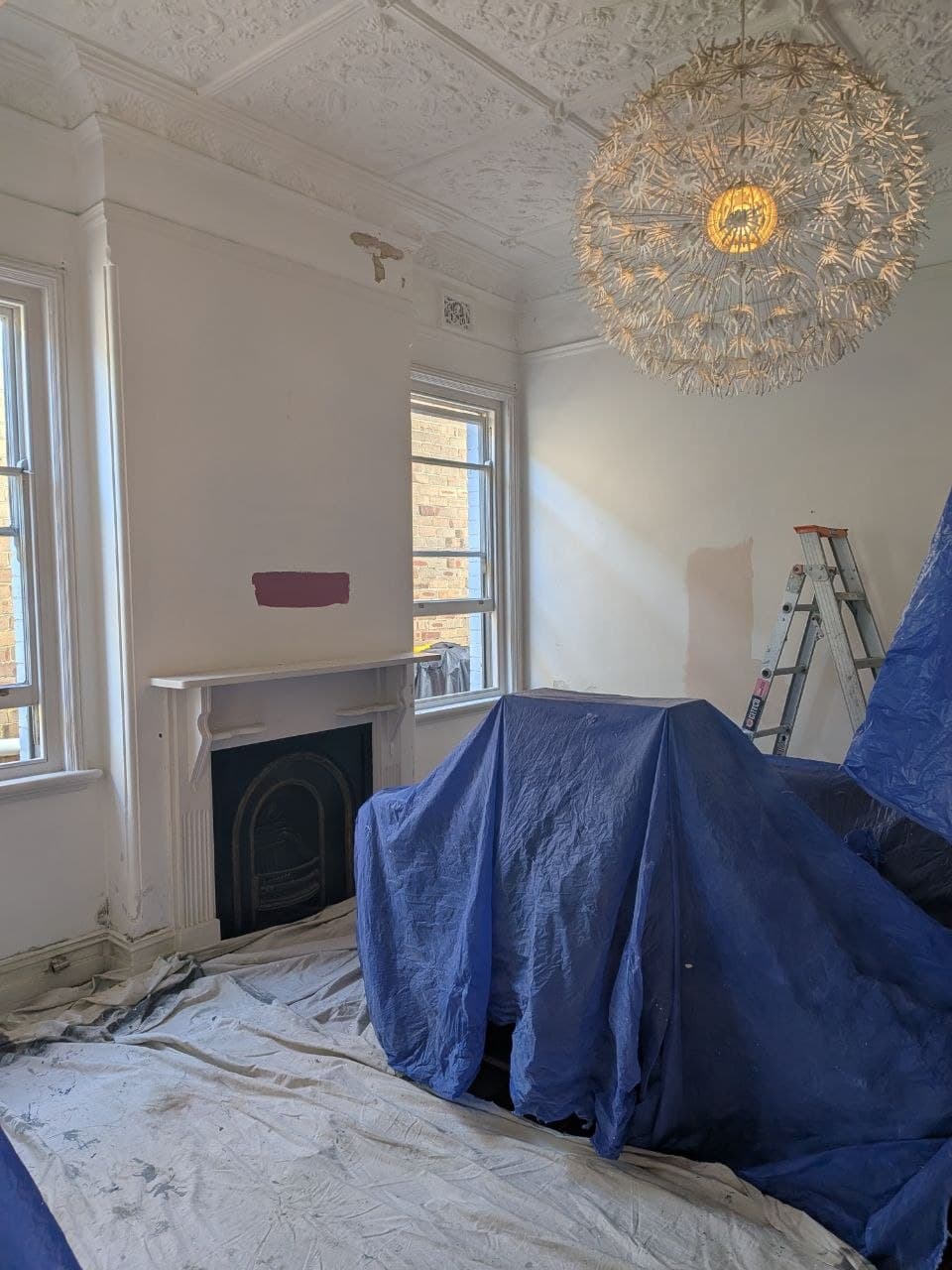 Living room mid-renovation — bare white walls, furniture under drop sheets and a ladder ready for painting